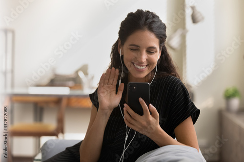Videocall. Happy millennial latin female in wired pair of headphones sit on couch talk to friend in video app wave hand say hi to caller. Friendly young lady use cell for video call make hello gesture