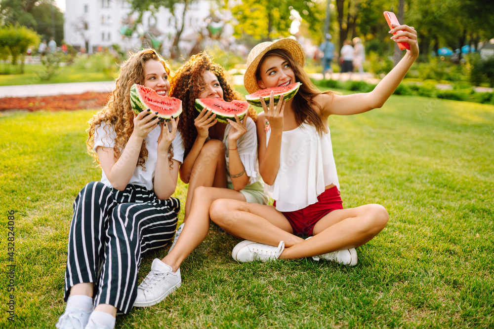 Fototapeta premium Three young woman relaxing on the grass, eating watermelon and taking selfie on the phone. People, lifestyle, travel, nature and vacations concept.