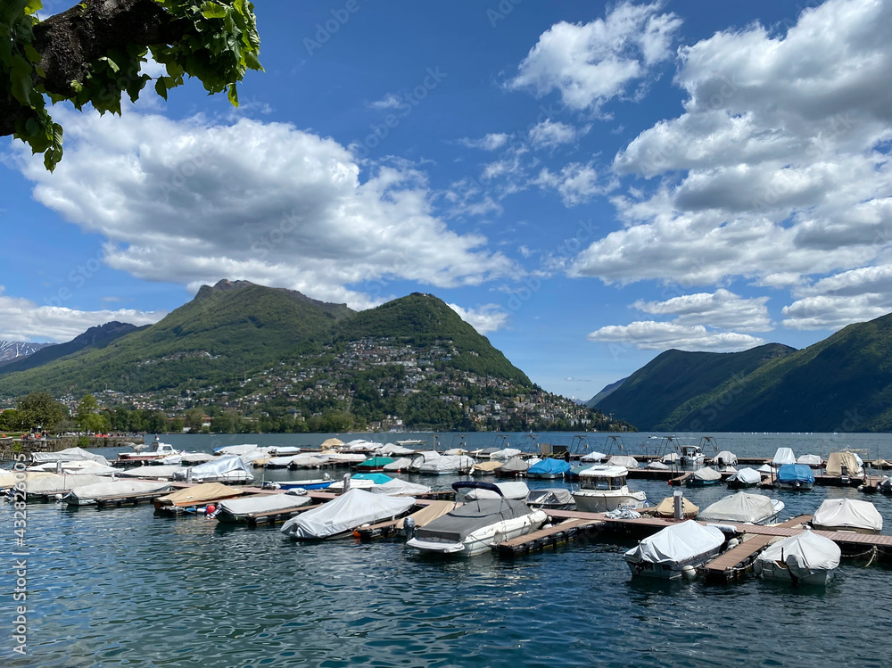 Lake Lugano. Switzerland. City center, embankment. View of the lake and mountains. Parked boats near the shore. Sunny day, clouds. Wallpaper Lake Lugano. Switzerland. City center, embankment. View of the lake and mountains. Parked boats near the shore. Sunny day, clouds. Wallpaper