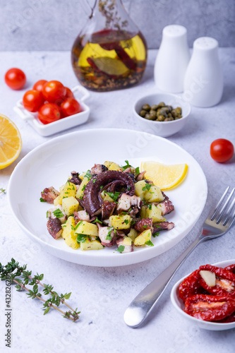 Warm salad with octopus, potatoes, tomatoes, capers and lemon on a white plate. Close-up, white background.