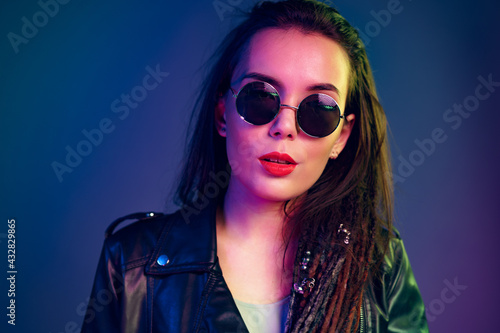 Asian girl in sunglasses and black leather jacket posing against dark blue background