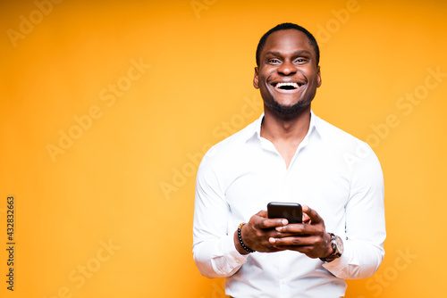 Happy african american man standing on yellow background and holding a phone in his hands © DmitryStock