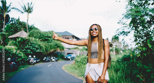 Cheerful African American hitchhiker with hand up catching automobile transport at roadside for getting to travel destination during wildlife style of trip, joyful female tourist enjoying vacations