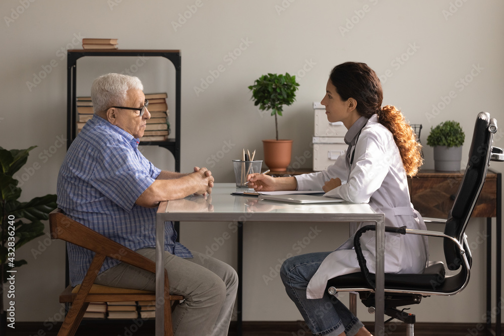 Obraz premium What is your complaint. Side view of older man patient meeting young woman attending physician for regular medical checkup. Senior male clinic visitor talking with latina female doctor at appointment