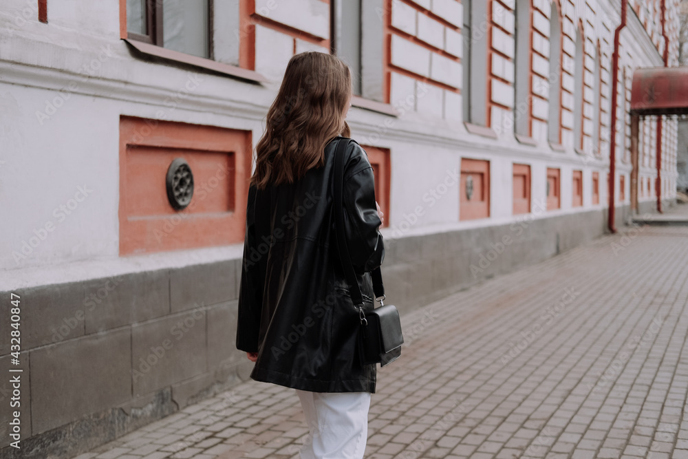 Unrecognizable woman in black leather jacket and white jeans walking on the street in front of white wall. Urban style and street fashion. Girl with curly hairstyle in casual outfit Unrecognizable woman in black leather jacket and white jeans walking on the street in front of white wall. Urban style and street fashion. Girl with curly hairstyle in casual outfit