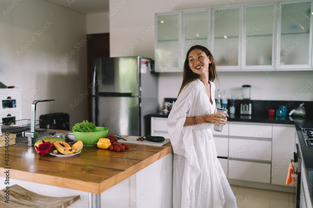 Positive young ethnic lady standing in stylish kitchen with water glass Positive young ethnic lady standing in stylish kitchen with water glass