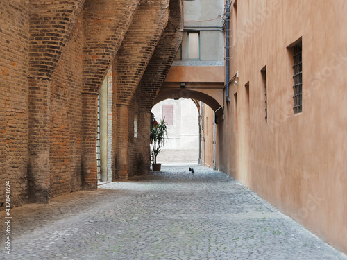 Fototapeta Naklejka Na Ścianę i Meble -  Ferrara, Italy. Closed-end alley next to the Municipal Theater. Cobbled street, on the left an old wall with buttresses. In the background you can see the Este castle.
