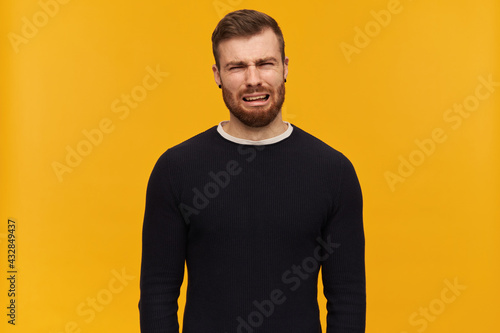 Bearded depressed guy, unhappy looking man with brunette hair. Has piercing. Wearing black sweater. Crying and wry face full of tears. Watching at the camera, isolated over yellow background