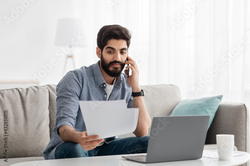 Arab freelancer man working remotely with smartphone, laptop and documents, sitting on sofa at home