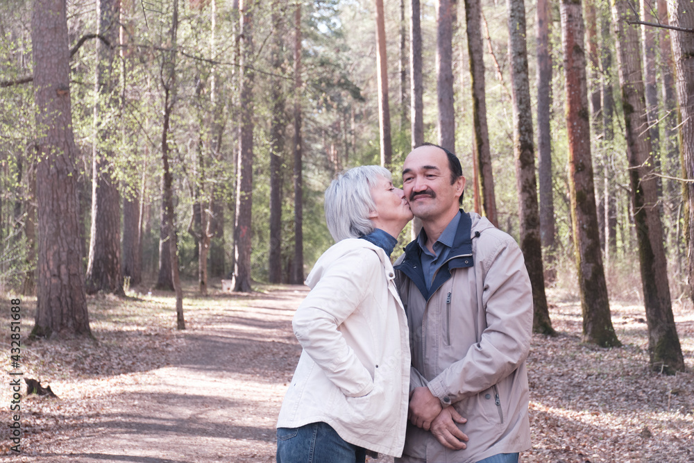 Elderly interracial couple in a spring forest park. A woman kissing a man on the cheek.
