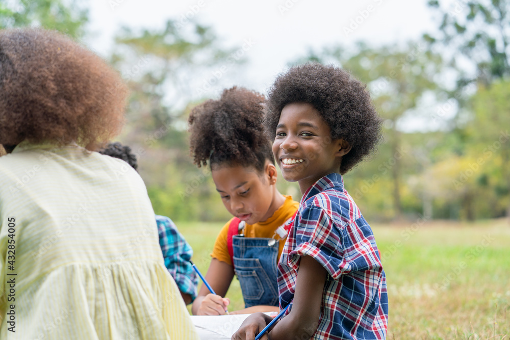Portrait of African American boy or Afro boy sitting with his friends on the grass having fun use pencils drawing on books in outside classrooms in the park of school. Education outdoor concept