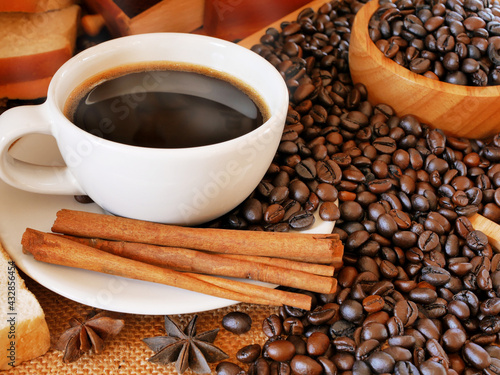 A white cup of aroma hot coffee near a wooden bowl of coffee beans and cinnamon for drink in the morning