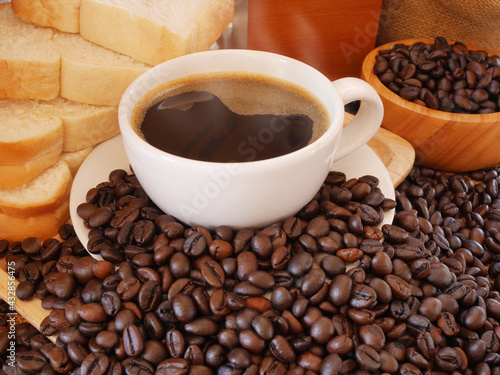 The cup of hot dark coffee is between more coffee beans and a wooden bowl of coffee beans and breads