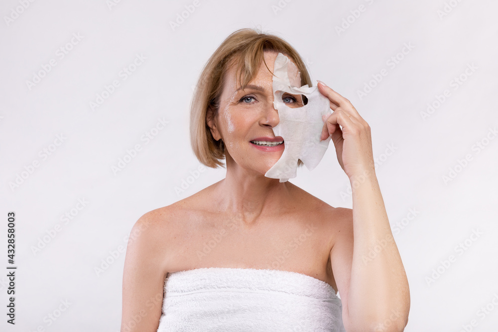 Portrait of happy senior woman in bath towel taking off rejuvenating cotton face mask on light studio background Portrait of happy senior woman in bath towel taking off rejuvenating cotton face mask on light studio background
