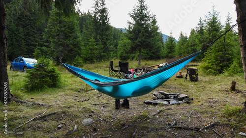 Sleeping in a hammock hanging between pine trees in a camping site in the middle of a coniferous forest. Some logs used as seats and table and a fireplace form a holiday landscape. Carpathia, Romania