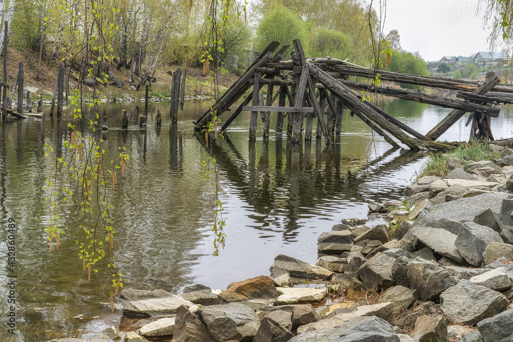 View of the old dilapidated wooden bridge over the river on a spring cloudy day. In the foreground, large stones lie on the shore, and a birch forest grows in the distance. Place near the village 