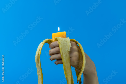 Mano di giovane ragazza tiene in pugno una banana sbucciata con all'interno una candela accesa dal basso su sfondo blu