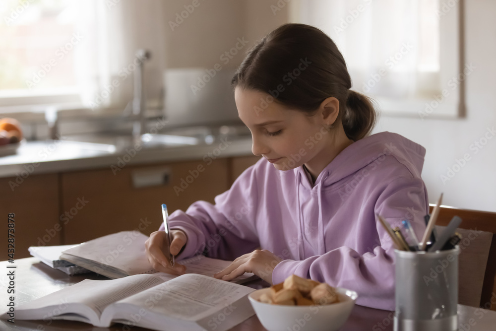 Focused adolescent kid girl sitting at table in kitchen, involved in doing homework or school assignment, preparing for examinations reading textbook, writing down important information in copybook. Focused adolescent kid girl sitting at table in kitchen, involved in doing homework or school assignment, preparing for examinations reading textbook, writing down important information in copybook.