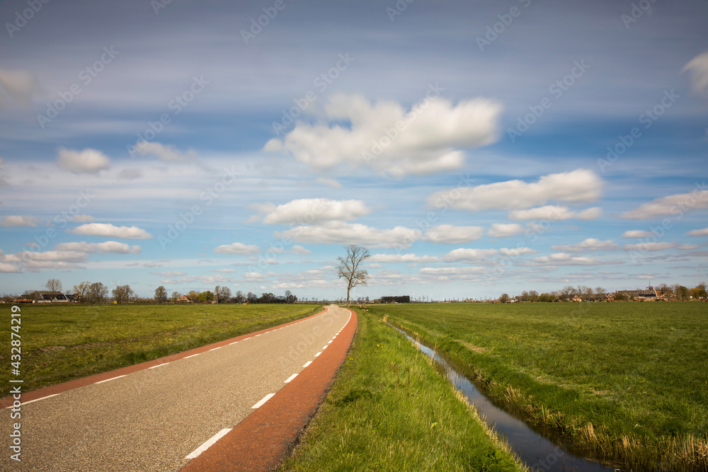Dutch landscape with a road, a cycling path, a tree, a canal with blue sky and clouds, province Groningen, the Netherlands