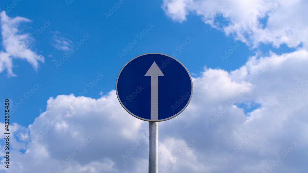 Round blue traffic sign with a white arrow among blue sky and white clouds Round blue traffic sign with a white arrow among blue sky and white clouds