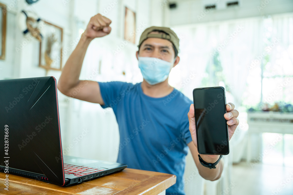 happy freelance asian young man wearing face mask working with laptop computer and show mobile phone with online job or shopping online happy and successful trading stock market in coffee shop cafe happy freelance asian young man wearing face mask working with laptop computer and show mobile phone with online job or shopping online happy and successful trading stock market in coffee shop cafe