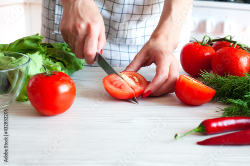 Womens hands with a knife cuts a fresh ripe tomato on wooden white board for salad. Healthy food concept. Vegetarianism. Close-up.