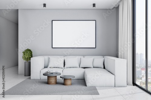 White poster with copyspace in black frame on light grey wall above big sofa in stylish spacious living room with plant in flower pot  coffee tables and city view from big window. 