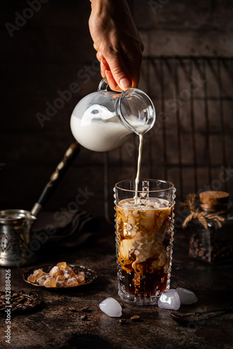 Ice coffee in a tall glass with cream poured over, sugar and coffee beans on dark background