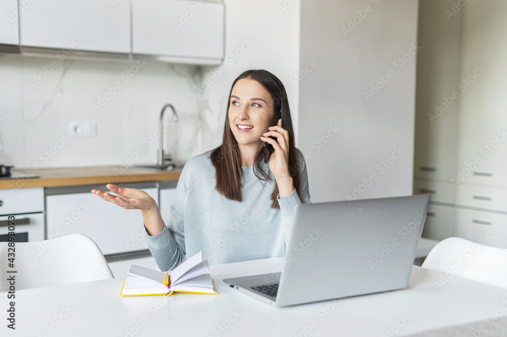 Smiling and friendly young woman talks on the smartphone using laptop computer at home, cheerful female holding phone call speaks with positive face expression, works remotely sitting in the kitchen