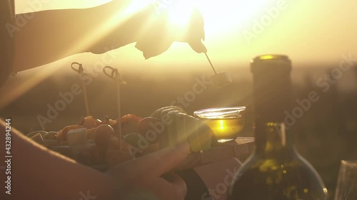 Against background of the bright sun, a woman's hand lowers a skewer of cheese (canapes) into plate of honey. a board with sliced fruit on the background of sunrise and sunset, Romance, bottle wine