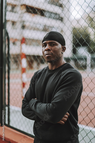 Confident Black Spanish male in black casual clothes and do-rag leaning on a wire fence photo