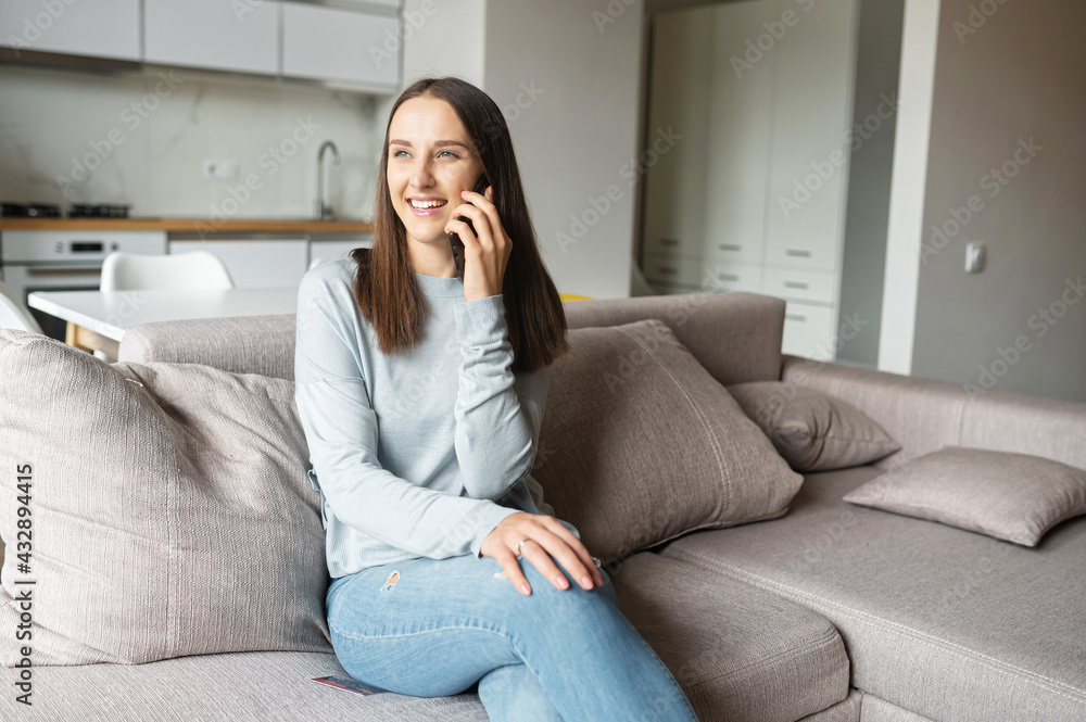 Serene young woman talks on the smartphone sitting on the couch at home, chatting with friends. Attractive female in casual wear sits on the comfortable couch and holding phone conversation Serene young woman talks on the smartphone sitting on the couch at home, chatting with friends. Attractive female in casual wear sits on the comfortable couch and holding phone conversation