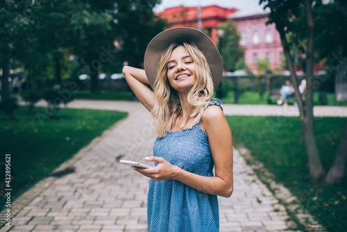 Happy female blogger in trendy hat holding modern cellphone technology enjoying summer daytime in park, cheerful Caucasian millennial woman with digital smartphone technology smiling outdoors