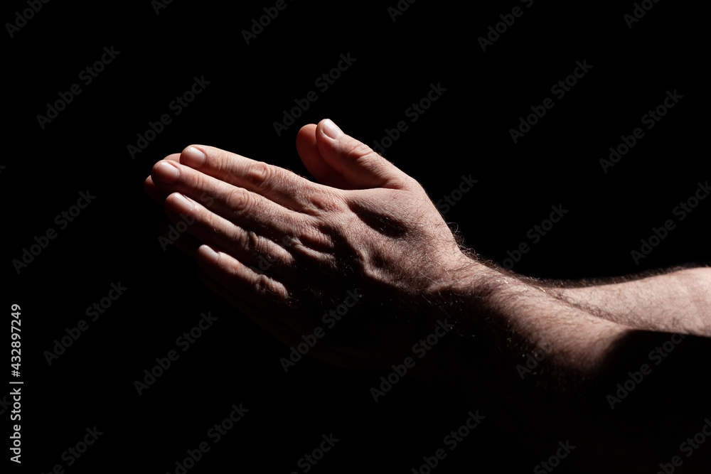 Fototapeta premium Male hands folded in prayer in the sunlight with harsh shadows, surrounded with darkness 