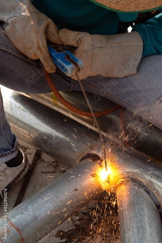 A welder is sitting on a steel roof frame and is welding steel pipes and wearing masks and leather gloves.