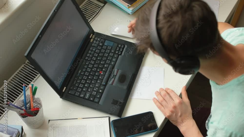 A student guy uses a laptop to study at home, he listens to words, makes notes on paper. Learning a new language. Top view. A student guy uses a laptop to study at home, he listens to words, makes notes on paper. Learning a new language. Top view.