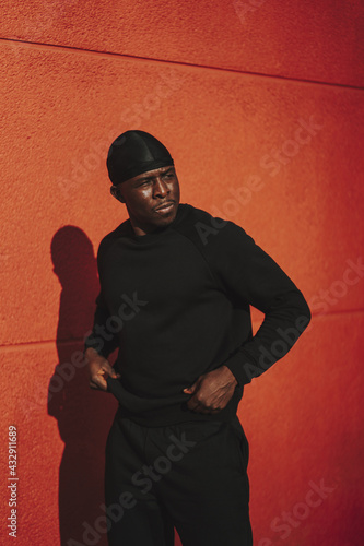 Confident Black Spanish male in black casual clothes and do-rag posing on a red wall background photo