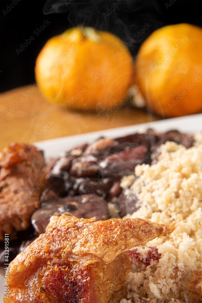 Brazilian feijoada, white dish with a delicious Brazilian feijoada, rice, farofa, and fried pork with two oranges on rustic wood, selective focus. Brazilian feijoada, white dish with a delicious Brazilian feijoada, rice, farofa, and fried pork with two oranges on rustic wood, selective focus.