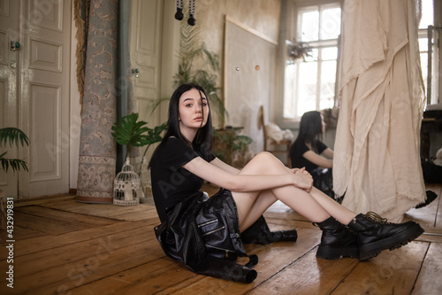 a beautiful brunette girl in a yodi and a black leather jacket sits on a wooden floor, a black thin pigtail on her face, big black ankle boots, reflection in the mirror, plants in pots