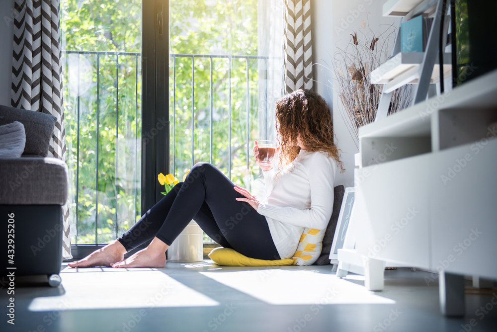 young woman sitting on the floor having coffee and looking out the window at home young woman sitting on the floor having coffee and looking out the window at home