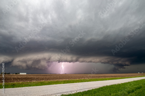 Lightning strikes as a supercell thunderstorm approaches a rural road. A shelf cloud marks the edge of the storm.