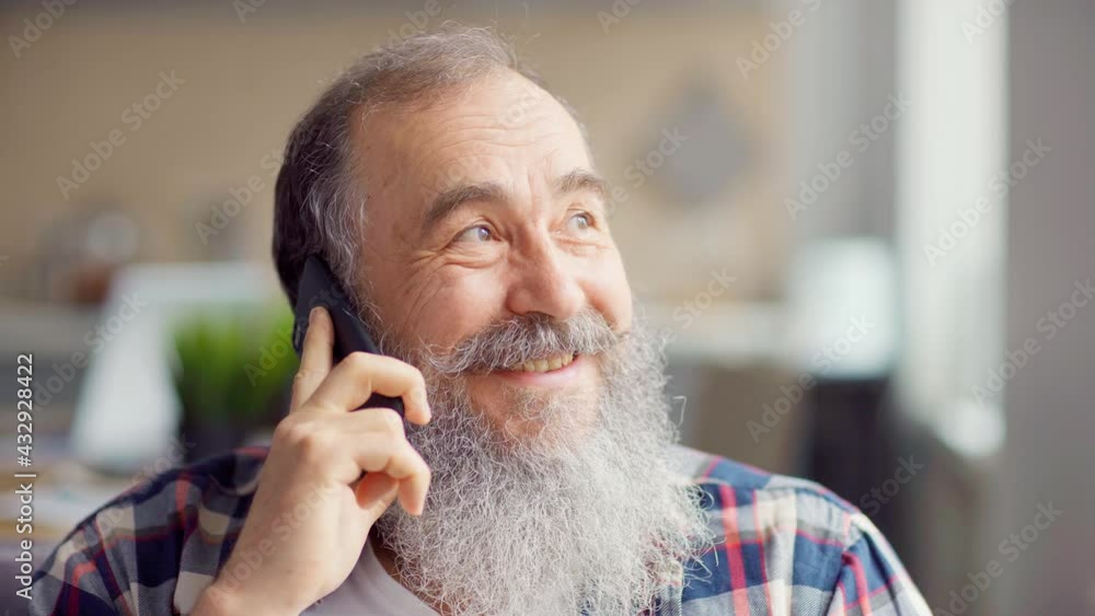 Head and shoulders shot of cheerful senior man with gray beard talking on cell phone at home, finishing call and looking at camera