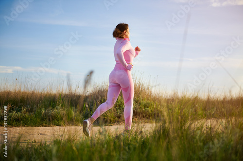 Young female athlete in pink sports outfit jogging on the steppe road with beautiful sunlight before the sunset