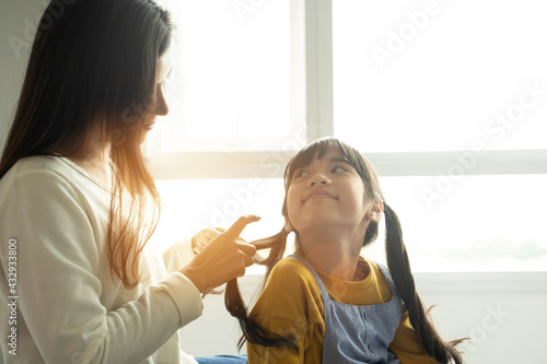 Asian mom making hair braid for daughter while sitting on bed at home.