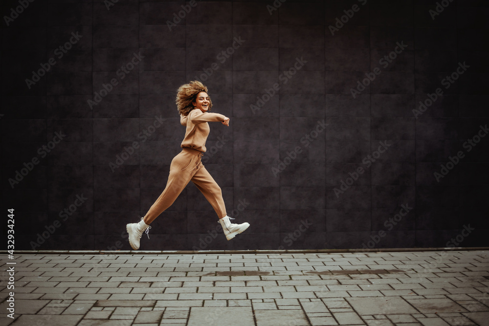 Smiling trap girl in tracksuit running next to a gray wall. - obrazy, fototapety, plakaty 