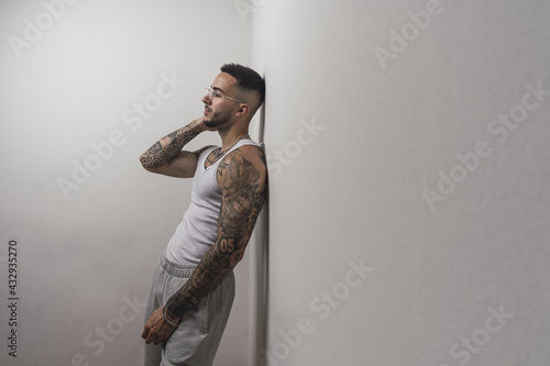 Portrait of a young handsome tattooed Spanish male posing leaning on a white wall