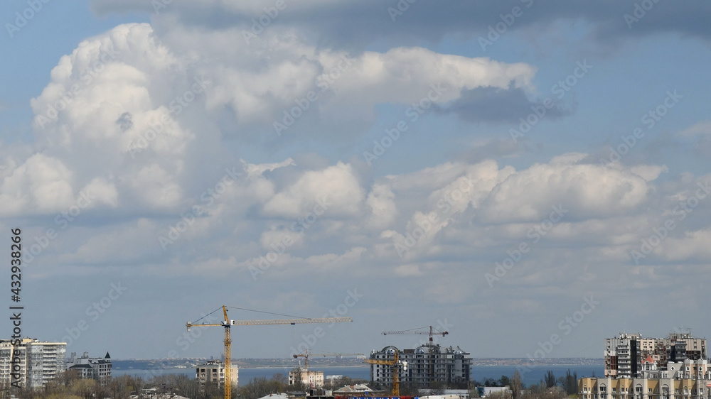 Construction crane jibs over rooftops under cloudscape. Under construction condo buildings in European city. Heavy construction machinery at urban skyline with cumulus clouds Construction crane jibs over rooftops under cloudscape. Under construction condo buildings in European city. Heavy construction machinery at urban skyline with cumulus clouds
