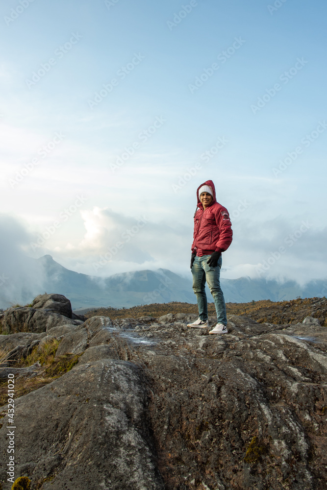 Hombre joven parado en la cima de una montaña mirando el paisaje con la nubes azules de fondo