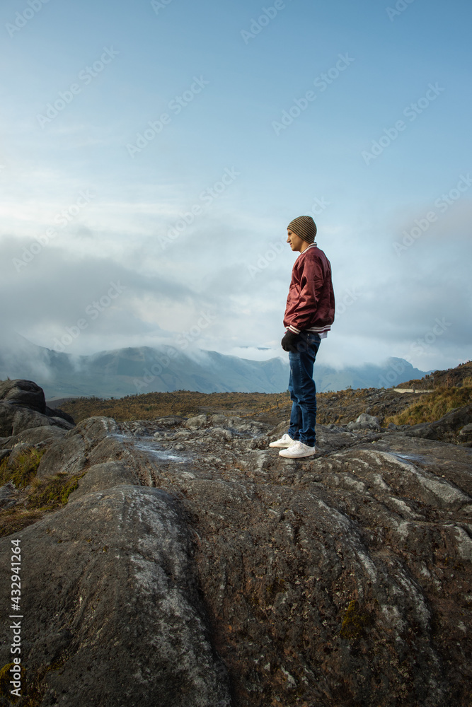 Hombre joven parado en la cima de una montaña mirando el paisaje con la nubes azules de fondo