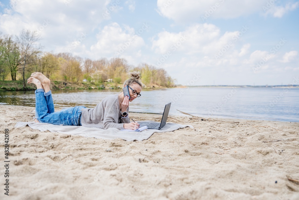 Blonde woman works outdoors on laptop. She's lying on the beach on a blanket and talking on the phone with klient.